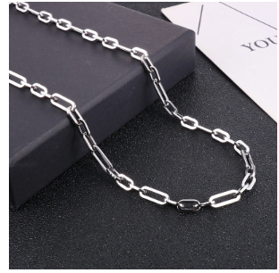 Collar de hombre en acero de titanio chapado en plata colocado sobre fondo negro, encima de una caja negra y junto a tarjeta blanca con la palabra “YOU”. Imagen de presentación que resalta calidad, acabado y estilo de la colección Hellochic@s-MEN Otoño-Invierno 2025/26