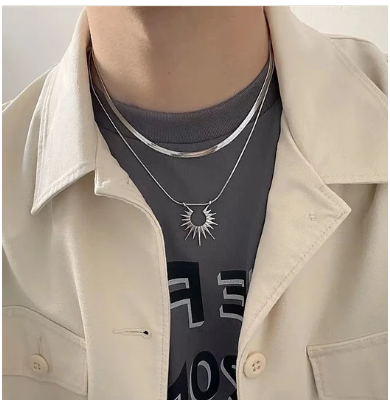 Modelo masculino vistiendo camiseta negra básica y camisa beige abierta, con collar de doble capa en acero de titanio chapado en oro 18K sobre la prenda oscura. La pieza resalta por contraste, mostrando versatilidad para looks casuales y urbanos. Colección Hellochic@s-MEN 2025/26